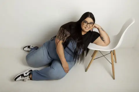 Smiling woman with long dark hair wearing glasses, a black t-shirt, and blue jeans sitting on a white floor.