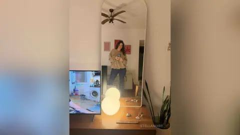 StellaNoire poses for a mirror selfie in a tan long-sleeve shirt and dark jeans in her living room.