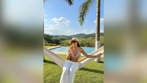 Smiling woman in white linen pants and bikini top relaxing in a hammock by a tropical pool on a sunny day.