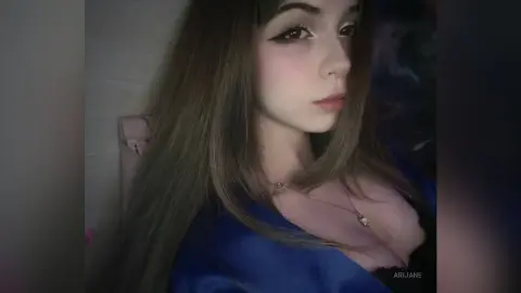 Young woman with long brown hair wearing a blue satin top in a moody indoor setting.