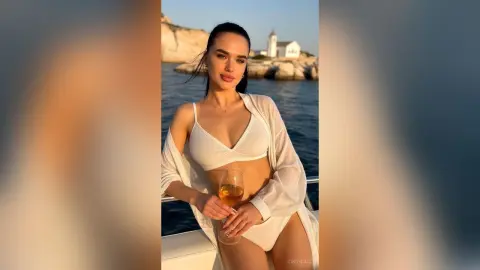 Young woman in a white bikini and sheer cover-up enjoying a drink by the ocean at sunset.