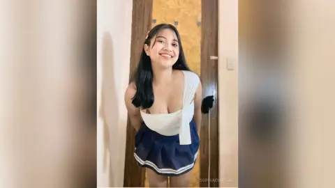 Smiling young woman in a white sleeveless top and navy pleated skirt posing in a bright indoor setting.