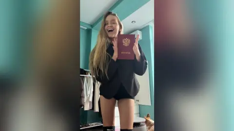 BonnyWalace smiles and holds up a red passport while wearing a black blazer and shorts in a teal room.