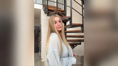 MeganPerson poses with her long blonde hair down, wearing a light blue button-down shirt while standing next to a wooden spiral staircase.