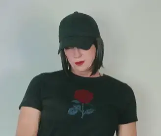 Young woman in a black rose graphic t-shirt and baseball cap posing against a plain wall with a moody vibe.