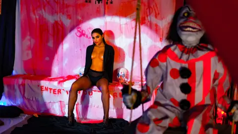 Woman in black lingerie sitting on a bed before a creepy clown in a spooky, red-lit horror room.