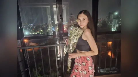 Young woman in a black crop top and Hello Kitty pajama pants holding flowers on a balcony at night.