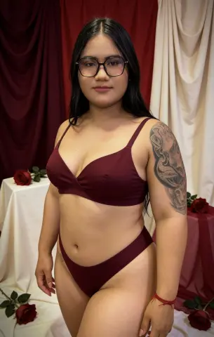 Lizzyolsen90 poses in a maroon bikini set, showing off her large shoulder tattoo and long black hair against a red and white curtain background.