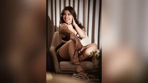 Smiling brunette woman in black lingerie sitting on a chair in a moody, dimly lit indoor setting.