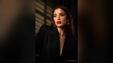 OliviaHardies poses in a black top with red lipstick and long dark hair in a dimly lit room.