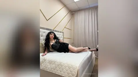 ErikaBradley lies on a white bed wearing a black tank top, showing her long dark hair and legs in a bright room.