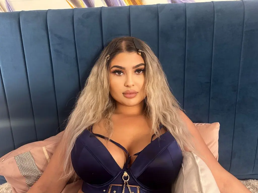 Blonde woman in a navy blue satin bodysuit relaxing against a teal velvet headboard in a cozy mood.