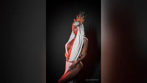 LeslyAnderson poses naked against a black background, wearing a white headwrap and a gold crown while holding her breasts.