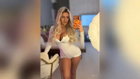 Anita poses in a white long-sleeved bodysuit, holding her phone for a mirror selfie in a bedroom setting.