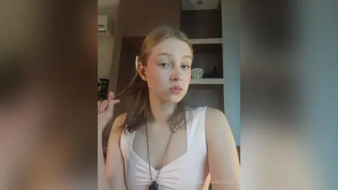 EvelinaMoorina looks into the camera wearing a white tank top and a necklace while touching her hair.