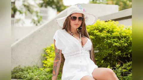 AndreaKalo wears a white sun hat and a white lace romper, showing off her long brown hair and colorful arm tattoo while sitting outdoors near green bushes.