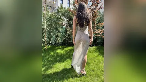 Woman in a long champagne satin dress standing in a sunny green garden setting.