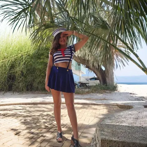 Smiling woman in navy sailor outfit posing under palm trees on a sunny tropical beach.