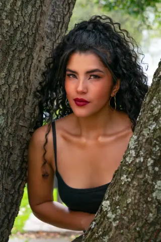 Young woman with curly hair in a black tank top leaning against a tree in a lush outdoor setting.