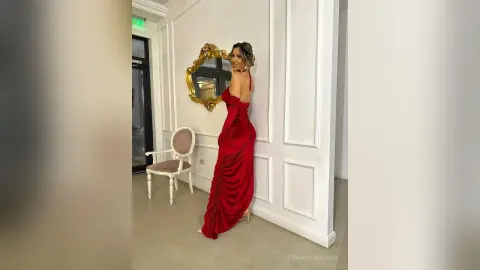 TiffanyAddams poses in a long, red, backless satin dress, looking over her shoulder in a white-walled room with a gold mirror and chair.