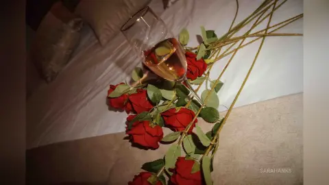 A glass of white wine resting among red roses on a white bedsheet in a romantic, moody setting.