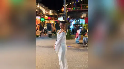 AnaMathers stands outdoors at night wearing a white off-the-shoulder long-sleeve top and white pants.
