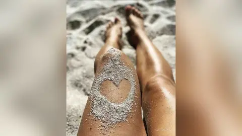 Close-up of a person's legs on a sandy beach with a heart shape drawn in the sand for a romantic summer mood.