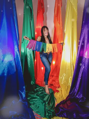 EmmaEbonyX19 smiles while holding a rainbow-colored fringe banner, wearing tight blue jeans and standing against a background of colorful satin drapes.