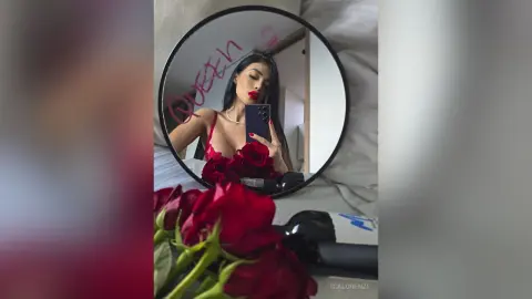 Glamorous woman in a red lace outfit taking a mirror selfie with red roses in a cozy bedroom setting.