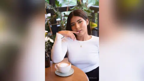 Young woman in a white long-sleeve shirt sitting at a cafe table with a latte in a bright, relaxed setting.