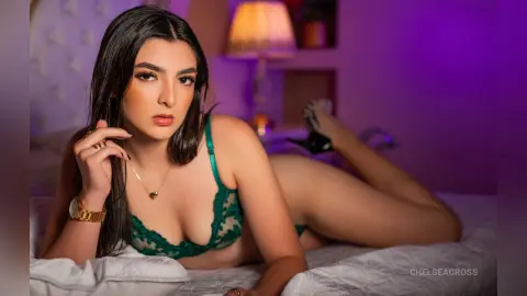 Young woman in green lace lingerie relaxing on a white bed in a moody, purple-lit bedroom setting.