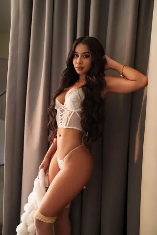 Beautiful woman in white lace lingerie posing elegantly against grey curtains in a soft, moody setting.
