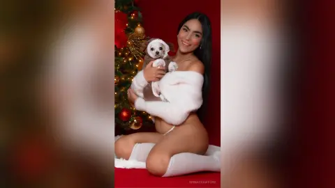 Smiling woman in white fuzzy loungewear hugging a small white dog by a festive Christmas tree.