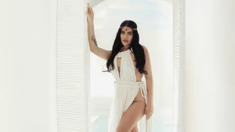 Elegant woman in a white Grecian goddess dress posing in a bright, airy Mediterranean villa setting.