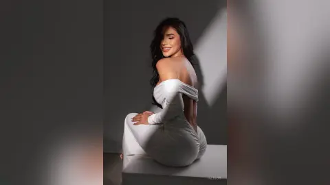 Alison poses sitting on a white platform wearing a white off-the-shoulder long-sleeve bodysuit that shows her bare back.