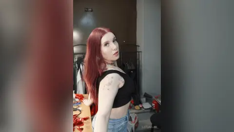 Red-haired woman in a black crop top and denim jeans posing in a modern minimalist bedroom.