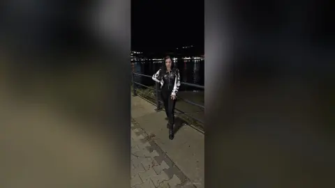 Stylish woman in a black and white patterned jacket standing on a waterfront walkway at night.