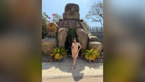 Stylish woman in a gold swimsuit posing in front of a large stone Buddha statue in a tropical garden setting.