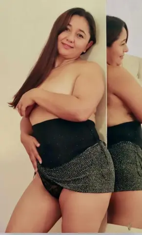 AngelRouss poses shirtless, wearing a black polka-dot skirt and black undies against a plain white wall.