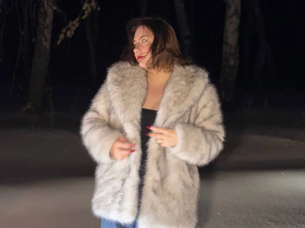 AnnyFraid wears a thick, light grey fur coat over a black top, posing outdoors at night with her hair down.