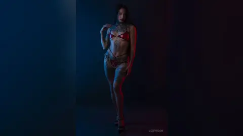 Tattooed woman in a red bikini posing confidently in a moody, dark studio setting.