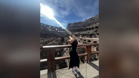Stylish woman in a black dress standing by the Colosseum ruins under a bright sunny sky in Rome.