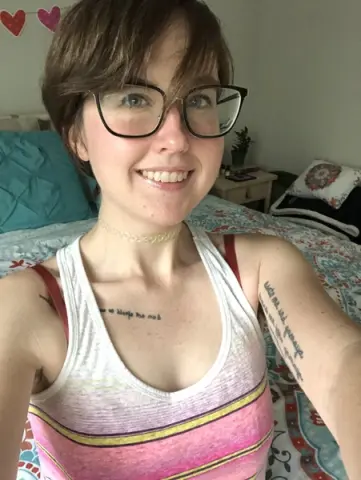 Smiling woman with glasses in a white tank top relaxing on a bed in a cozy bedroom setting.
