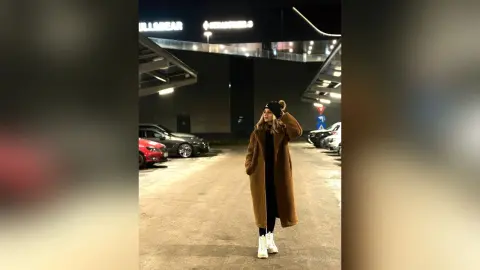 SofiaRoose stands outside at night wearing a long brown furry coat, a black beanie, and white boots.