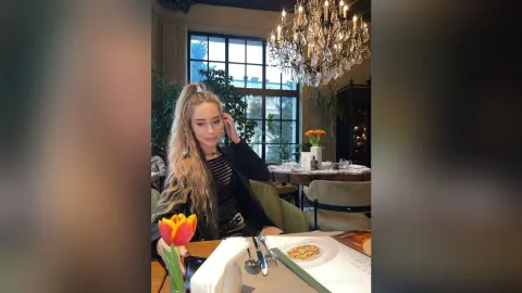 Blonde woman in a black outfit sitting at a dining table with flowers under a crystal chandelier.