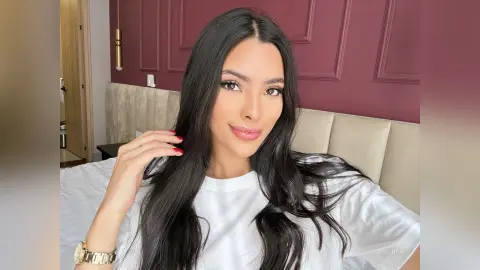 Smiling woman with long black hair wearing a white t-shirt in a modern bedroom setting.