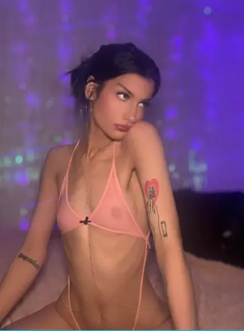 Young woman in a pink sheer bikini posing in a moody purple lit room with heart tattoos.