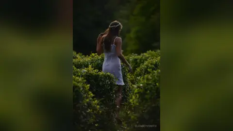 Dreamy woman in a white summer dress walking through a lush green garden at sunset.
