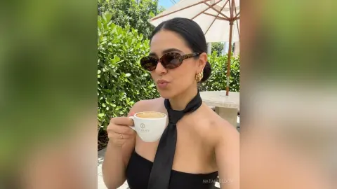 NatalyBloom wearing sunglasses and a black top, sipping coffee outdoors near green bushes.
