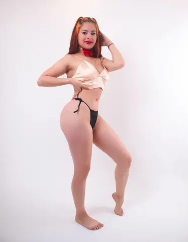 Redhead woman in a satin cream crop top and black bikini bottoms posing against a white studio background.